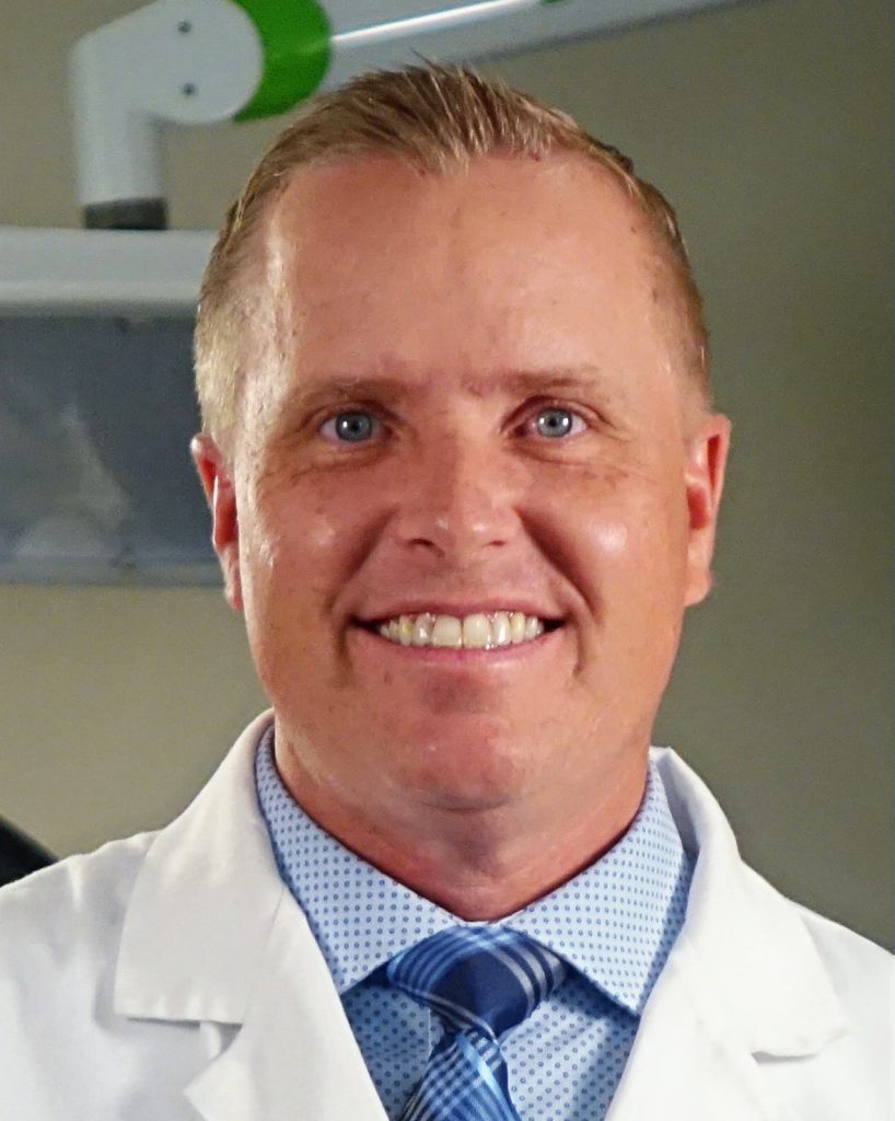 Dr. Brody J. Hart, DDS, smiling in a dental office, wearing a white coat and blue patterned shirt, representing high-quality dental care at Ivory Dental Implant Center.