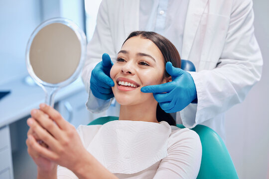 Woman in dental chair smiling while looking in a mirror, dentist adjusting her smile, showcasing satisfaction with dental care and single dental implants.
