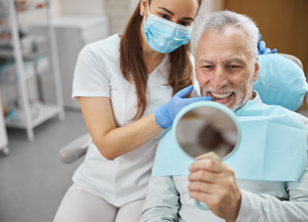 Patient smiling and holding a mirror, examining his dental results with a dental professional assisting, in a modern dental clinic setting, reflecting the expert care and satisfaction from dental implant services at Ivory Dental Implant Center.